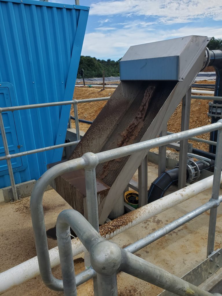 An inclined mesh screen filter at a palm oil wastewater plant, coated in reddish-brown solids strained from effluent, with a covered lagoon and blue equipment housing visible in the background.